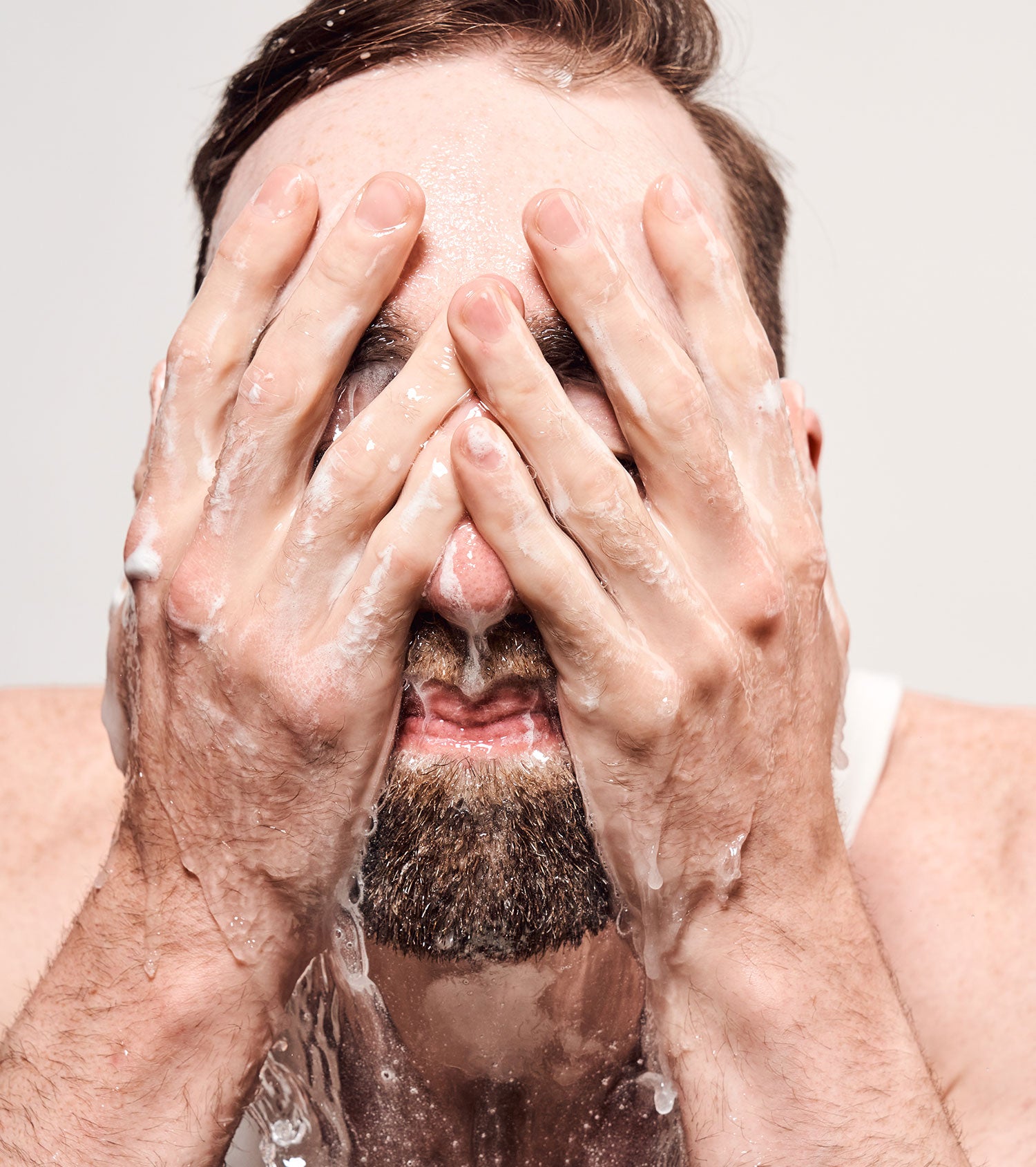 Man washing his face with soapy hands against a plain background