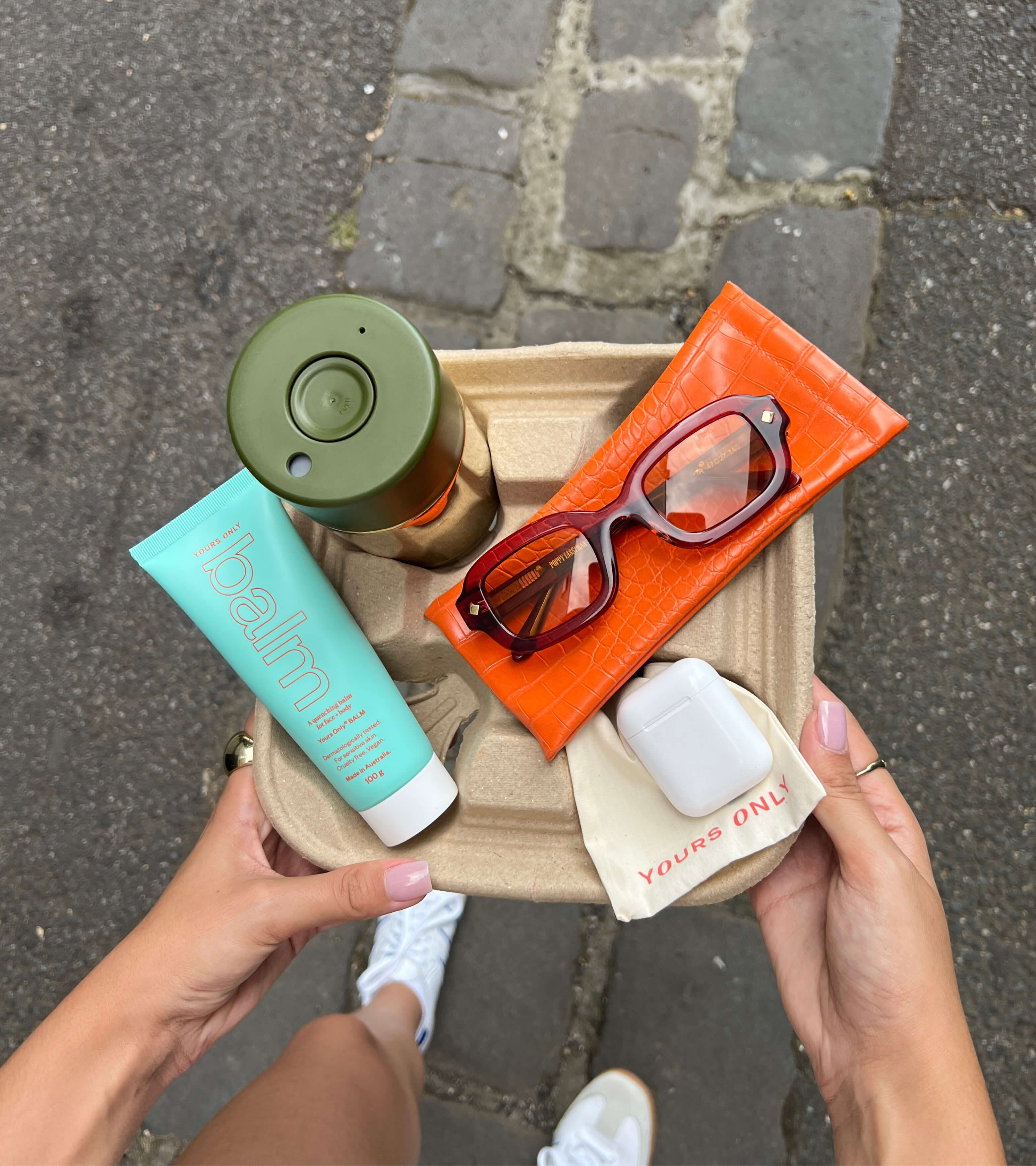 Hands holding a tray with lip + body balm, a green drink bottle, sunglasses, and wireless earbuds