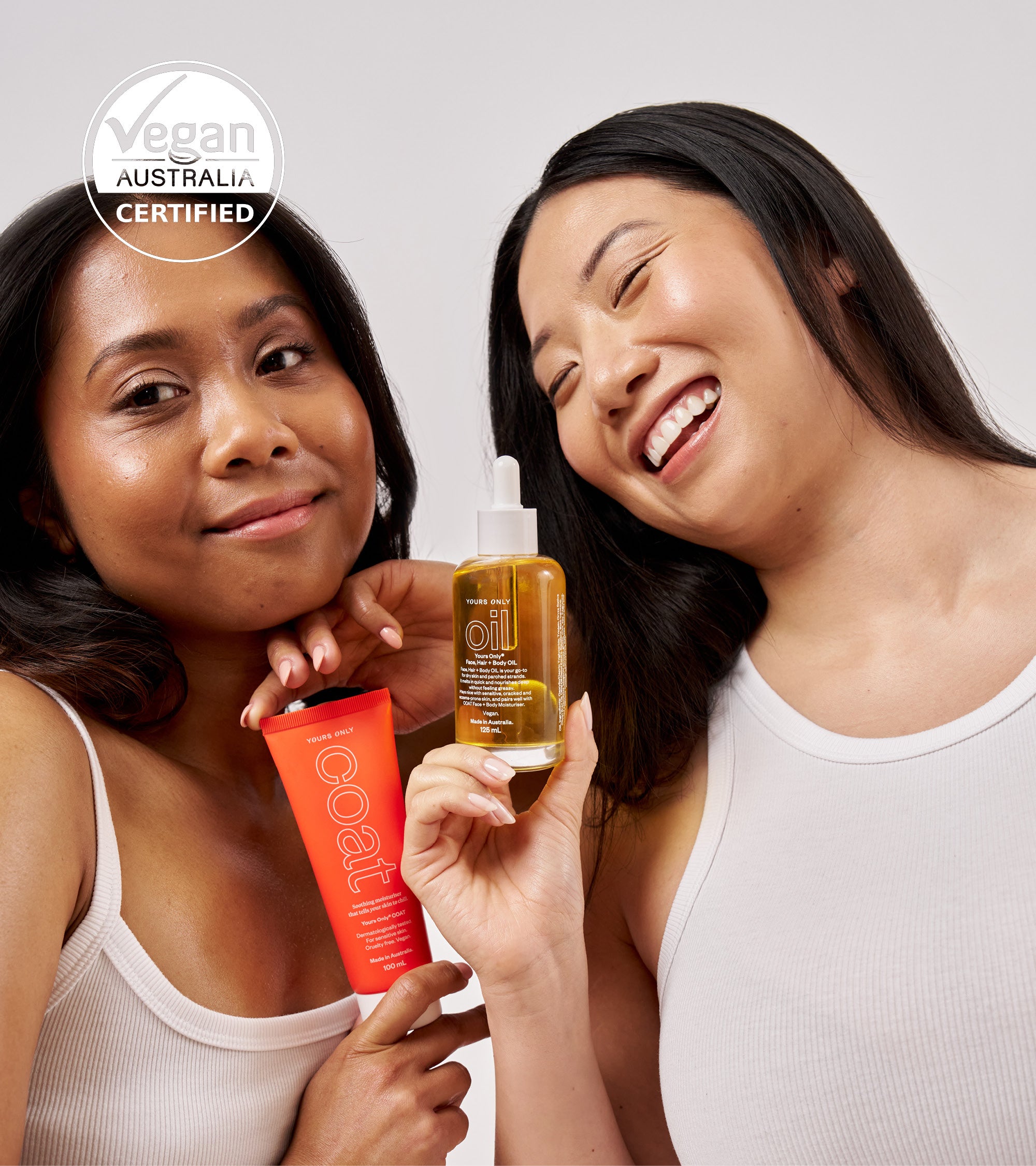 Two women holding a bottle of oil and a tube of product, with a 'Vegan Australia Certified' logo in the corner.
