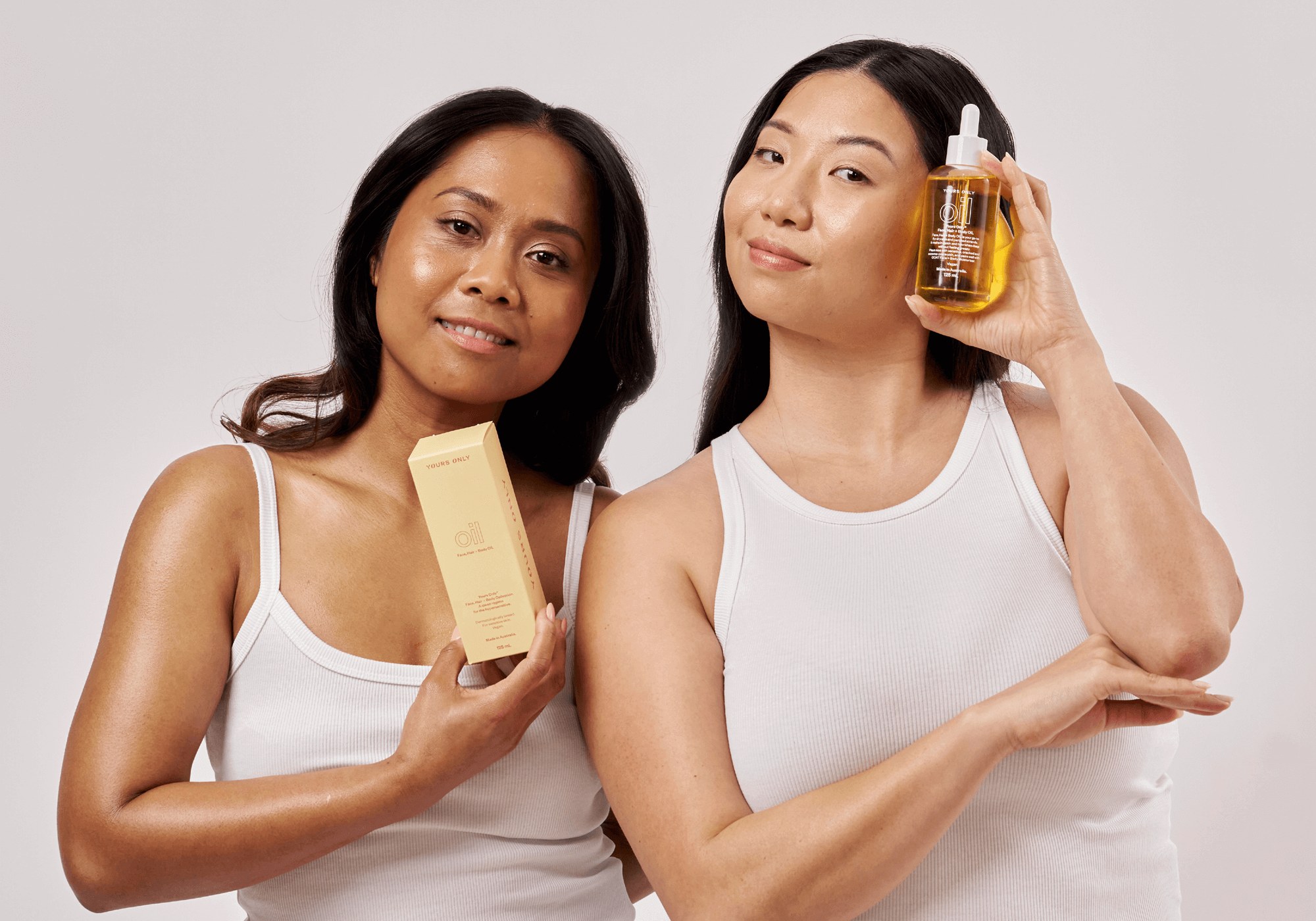 Two women in white tank tops holding Yours Only Face, Hair, Body Oil products